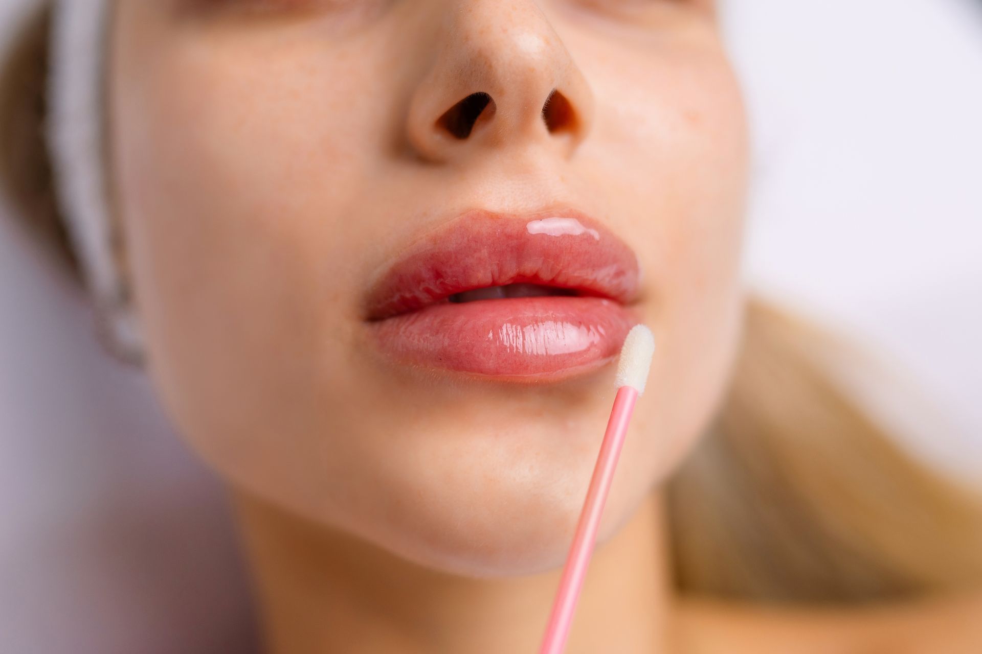 Woman with full, glossy lips being touched with a cotton swab, likely during a cosmetic procedure.