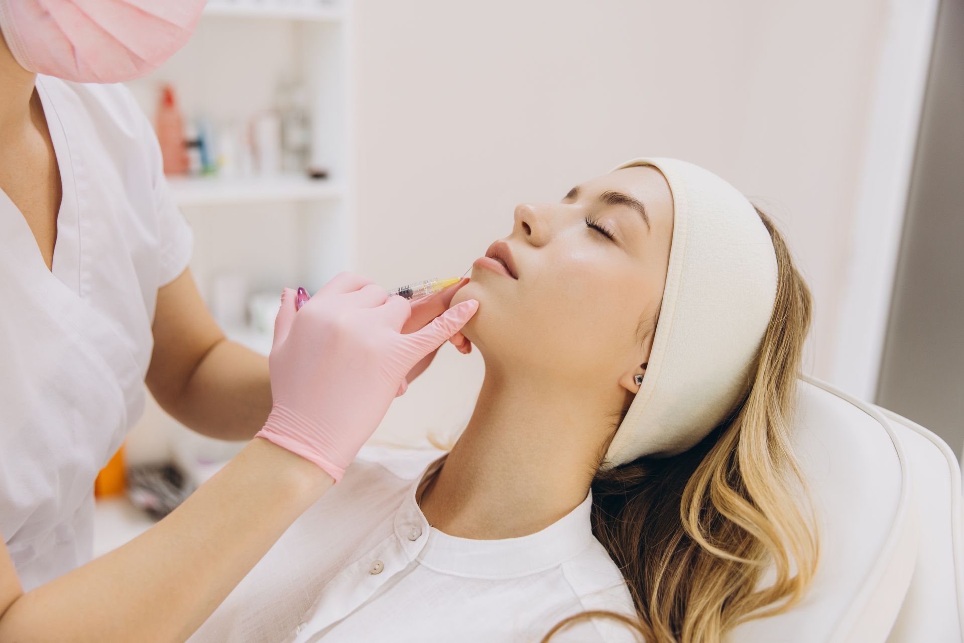 A woman receiving a facial injection. A healthcare professional in gloves and mask performs the procedure in a bright clinic.