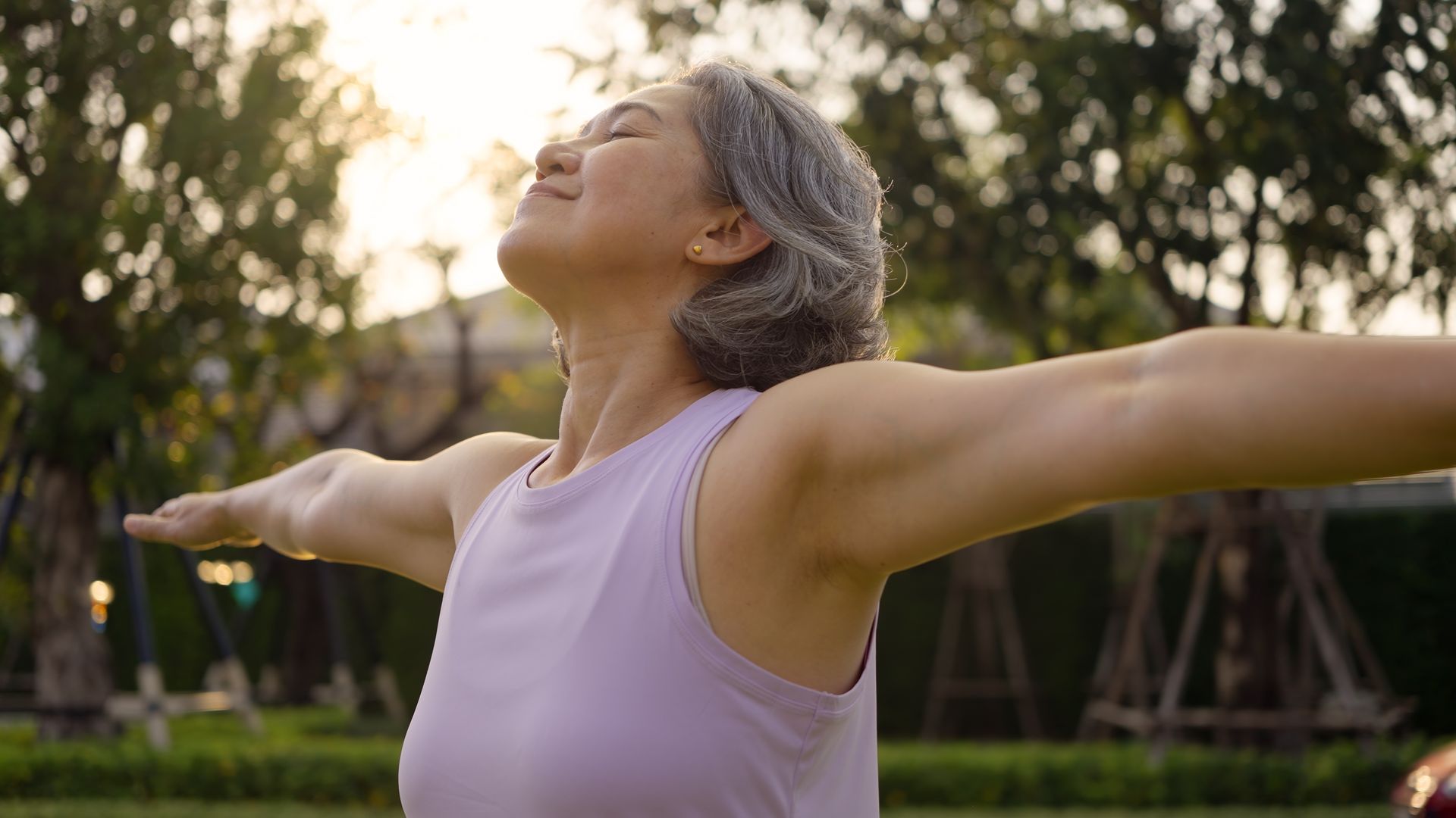 Woman with gray hair, arms outstretched, eyes closed, in a park, bathed in sunlight.