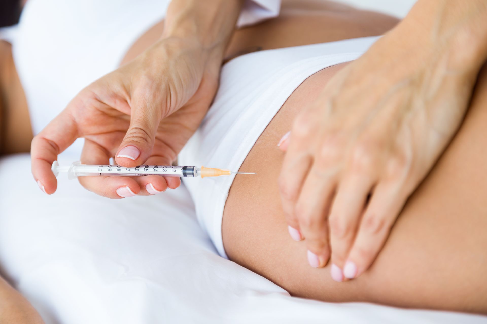 A person receiving an injection in their thigh; hands holding syringe, in a medical setting.
