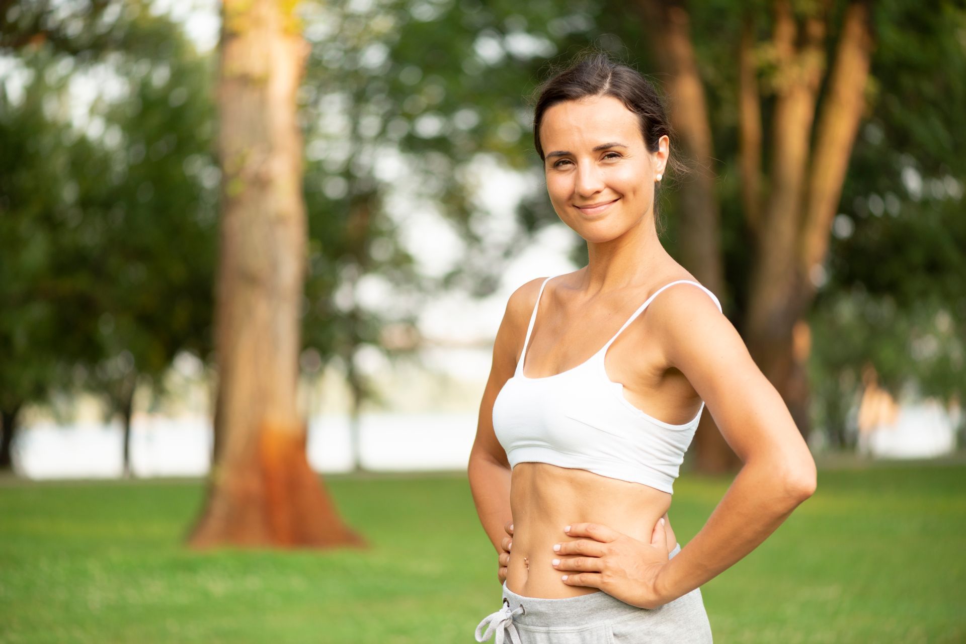 Woman in white sports bra and gray sweatpants smiles outdoors with hands on hips.