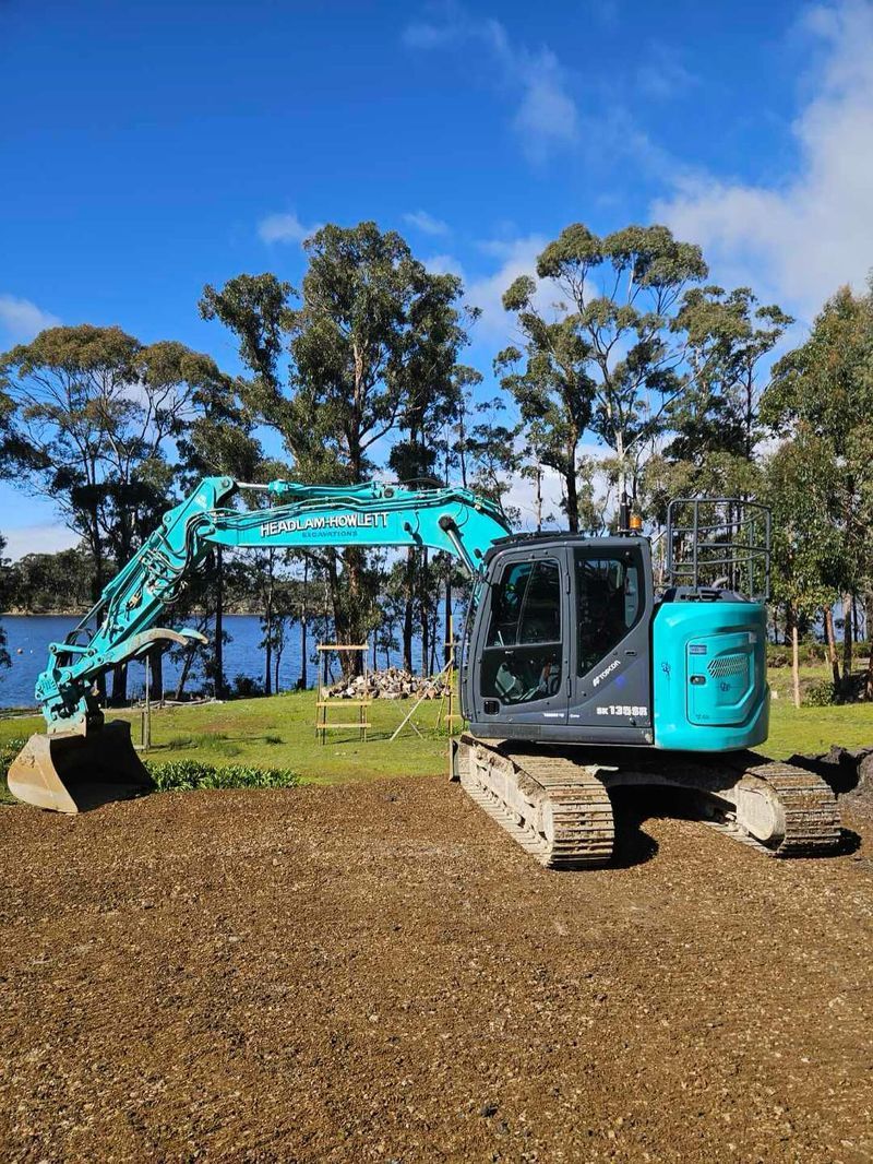 Blue excavator parked on brown dirt, overlooking a lake with trees under a blue sky.