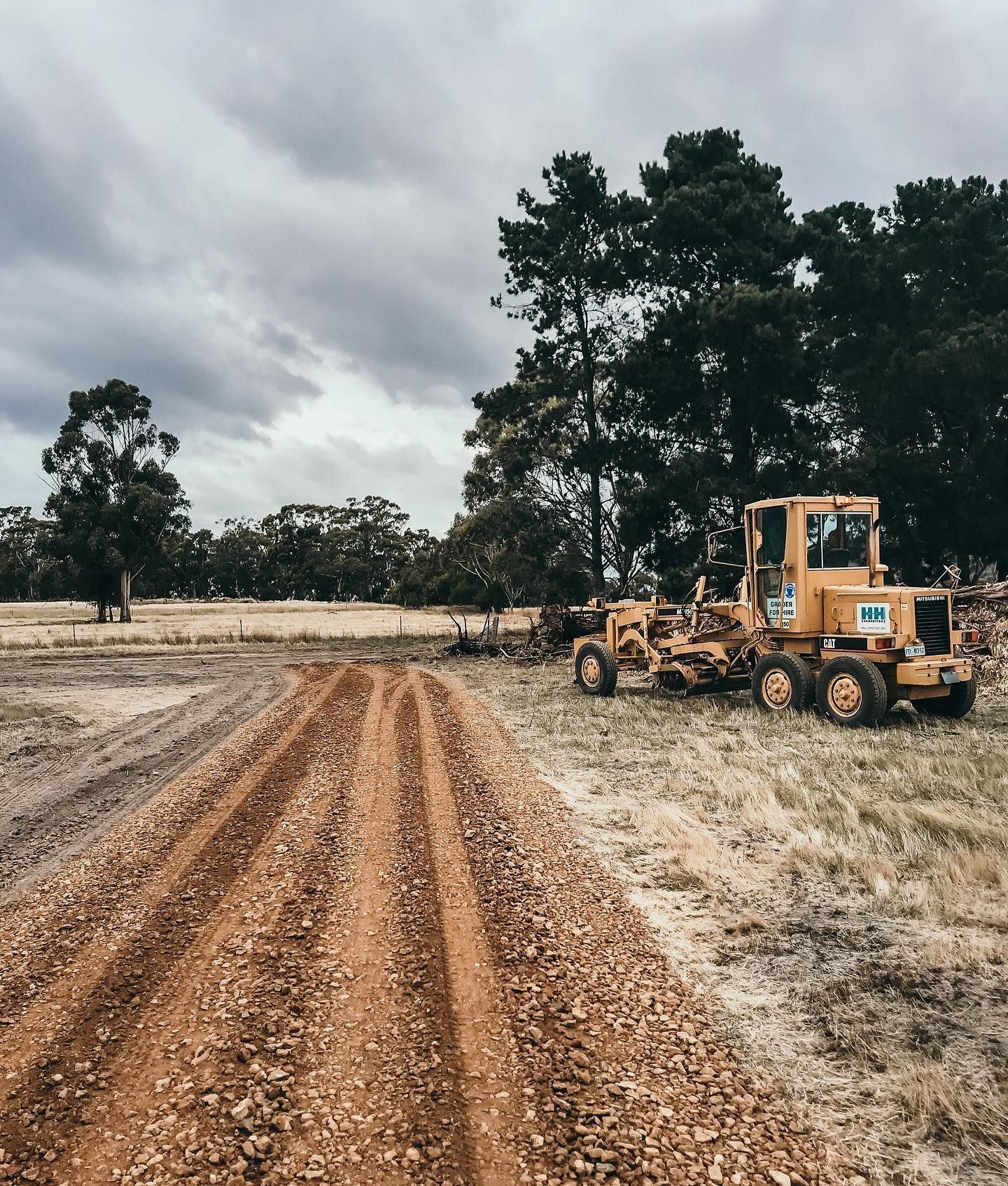 Yellow grader on a dirt road, trees and overcast sky in the background.