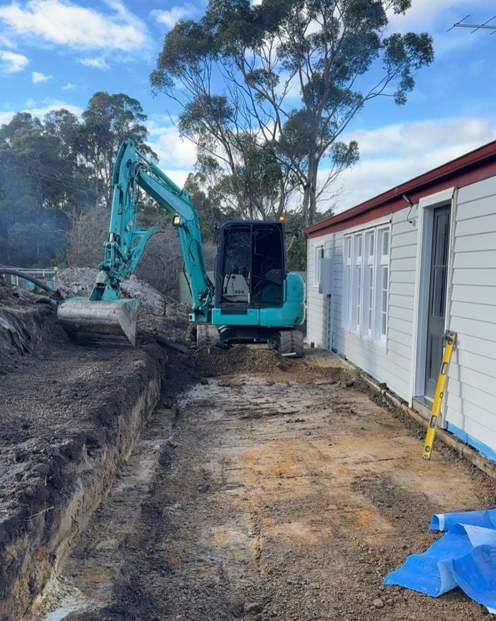 An excavator digs beside a white building. The sky is cloudy and trees are in the background.