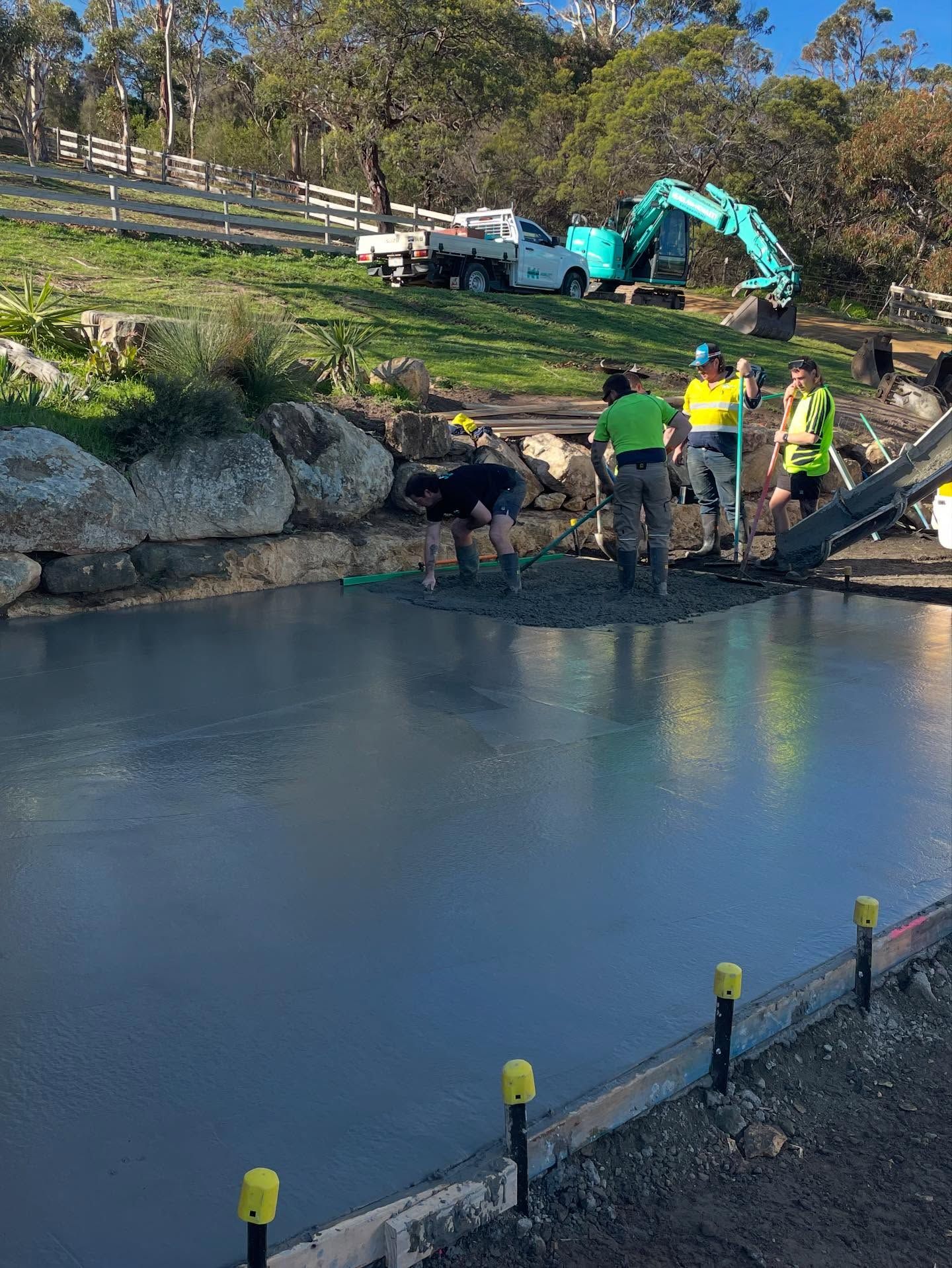 Construction workers pouring concrete on a flat surface, with an excavator and truck in the background.
