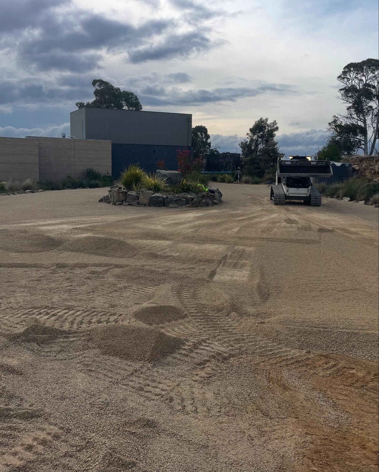 Gravel driveway with a truck, a low rock garden, and a modern building under a cloudy sky.