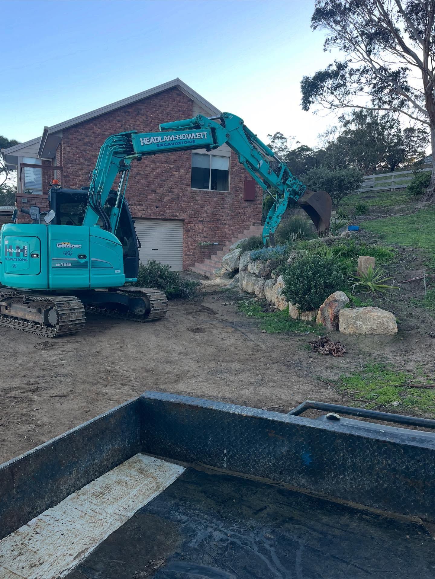 Teal excavator digging near a brick building. Dump truck in foreground.