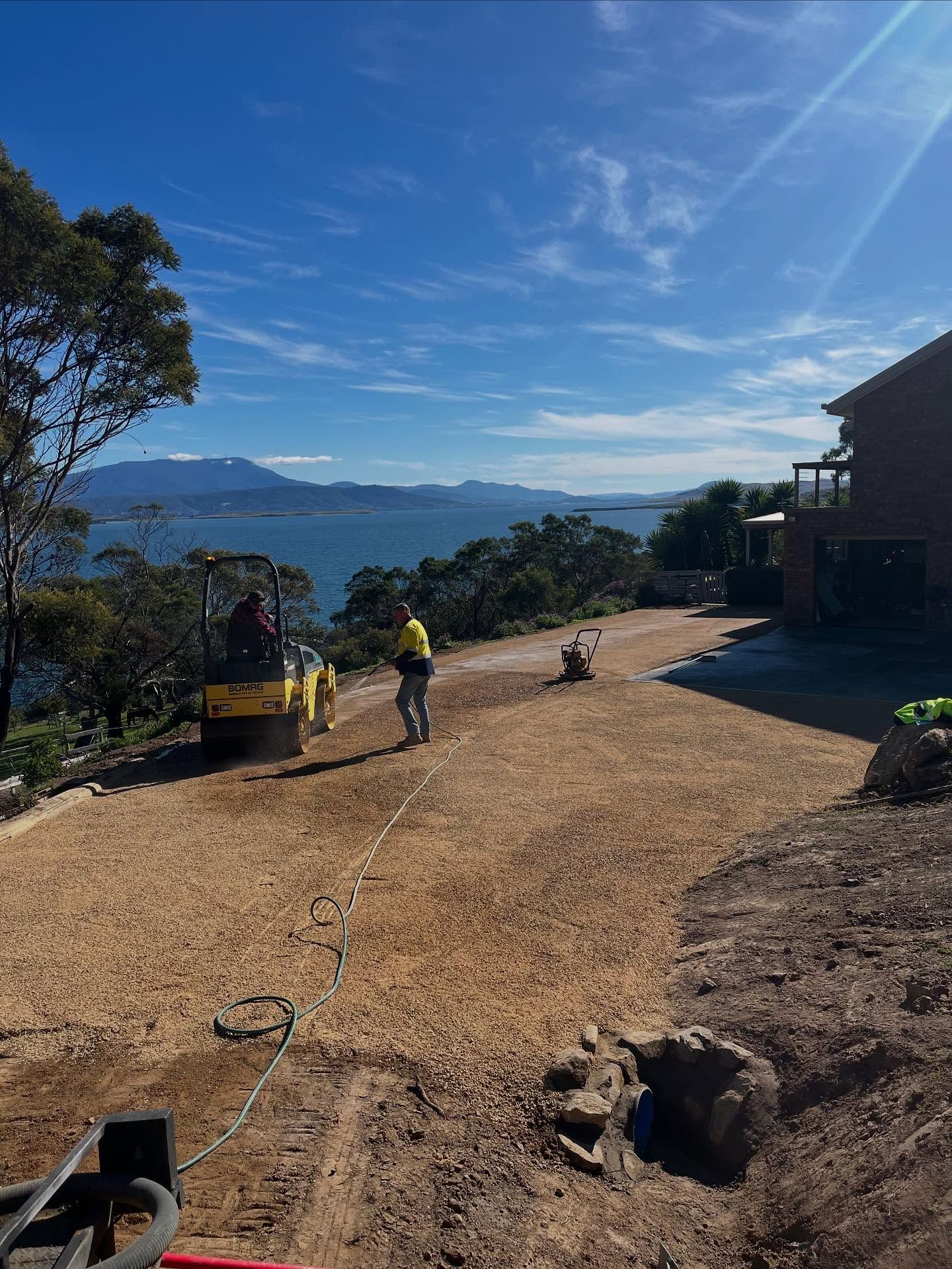 Construction workers compacting gravel driveway with a roller, scenic view of water and sky.