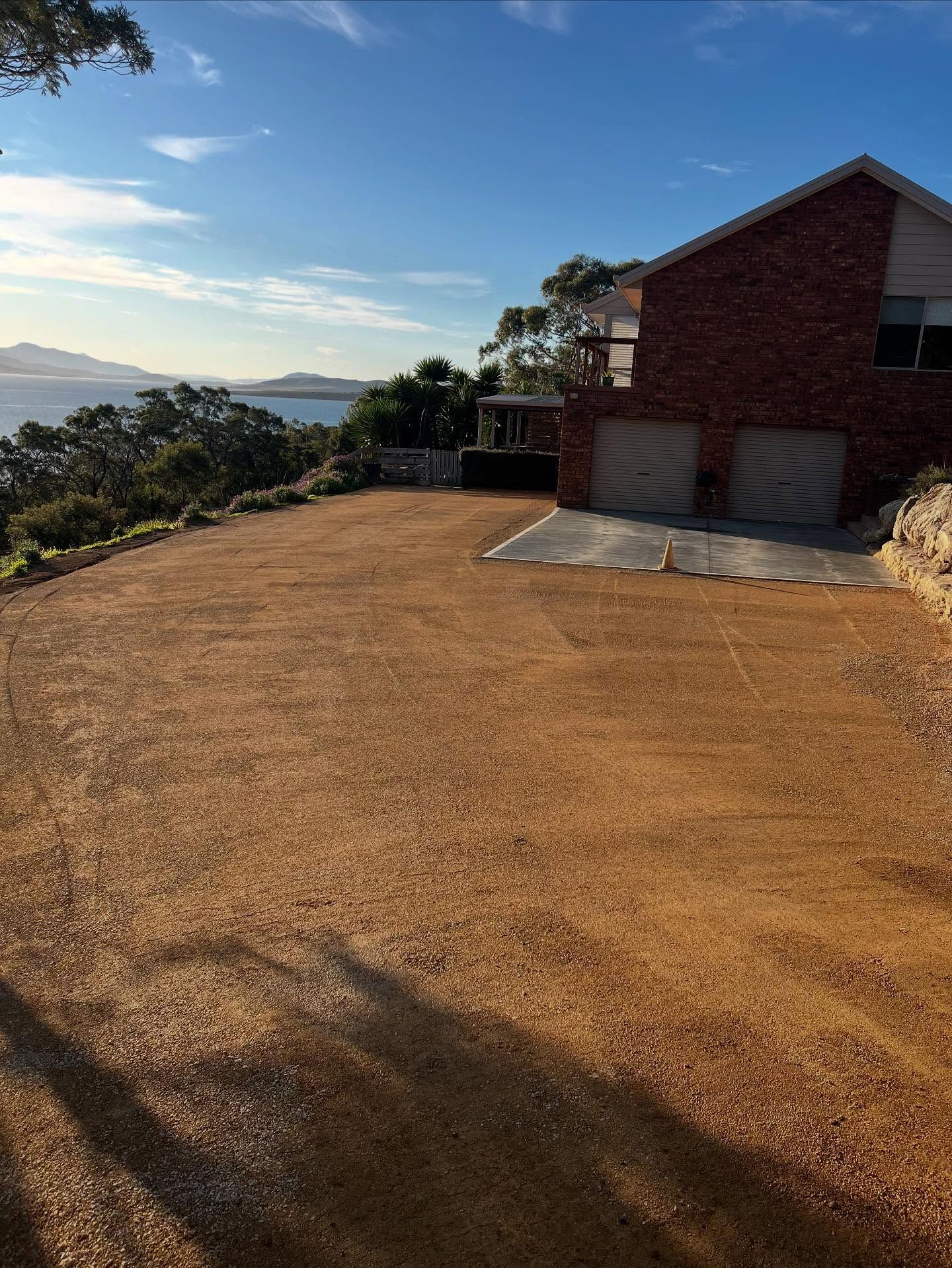 Gravel driveway in front of a brick house, overlooking a bay under a blue sky.