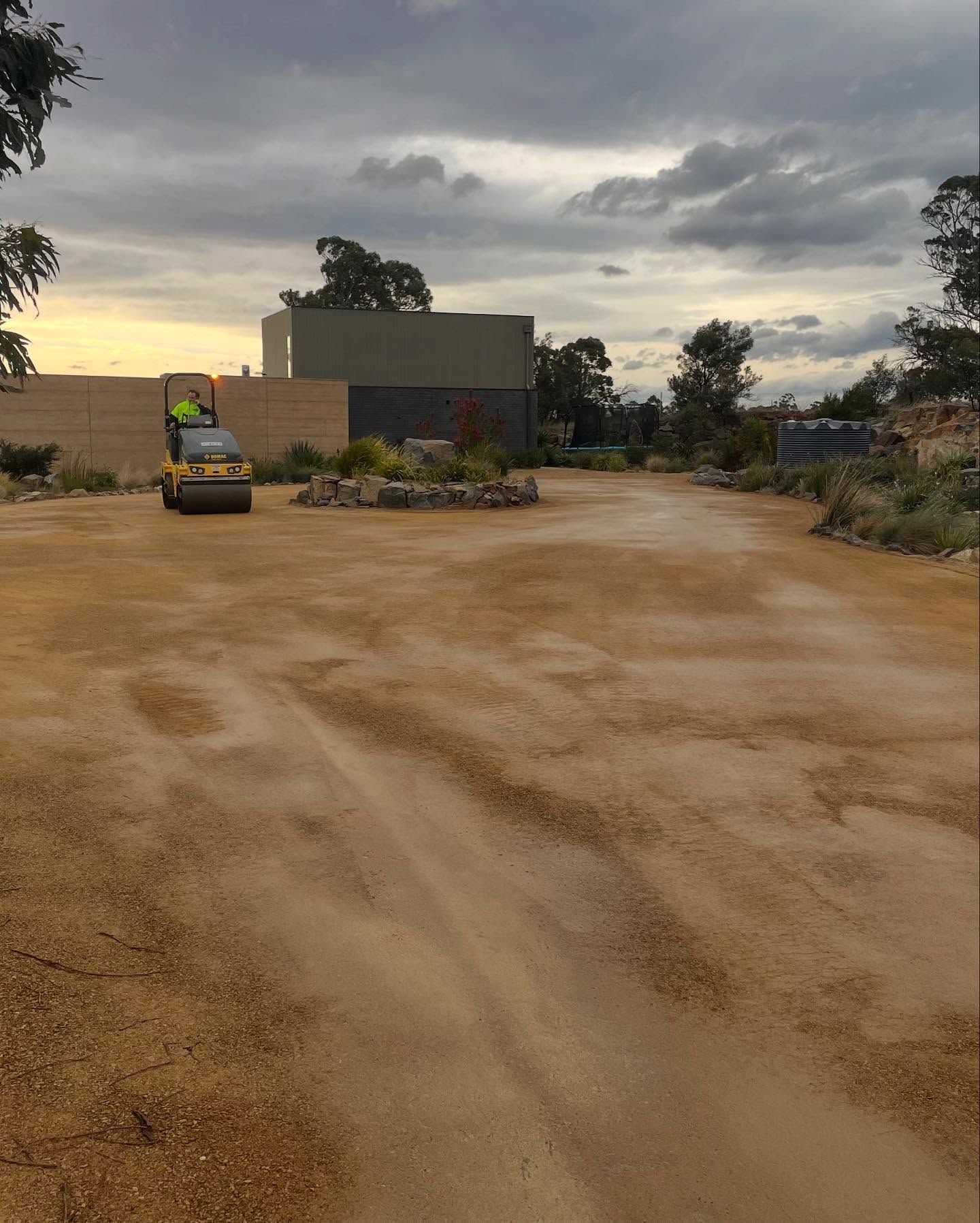 A worker operating a road roller compacting gravel in a construction area, cloudy sky background.