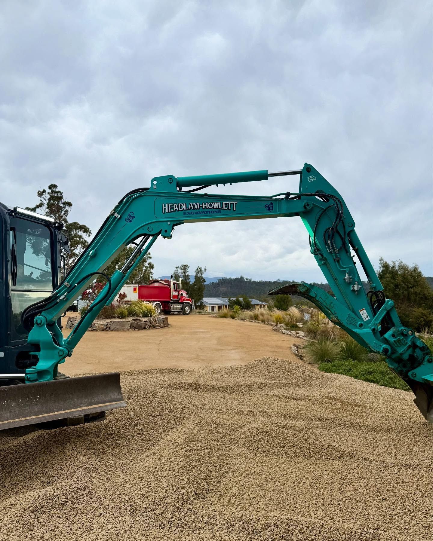 Green excavator arm on a gravel driveway, red truck in the background, cloudy sky.