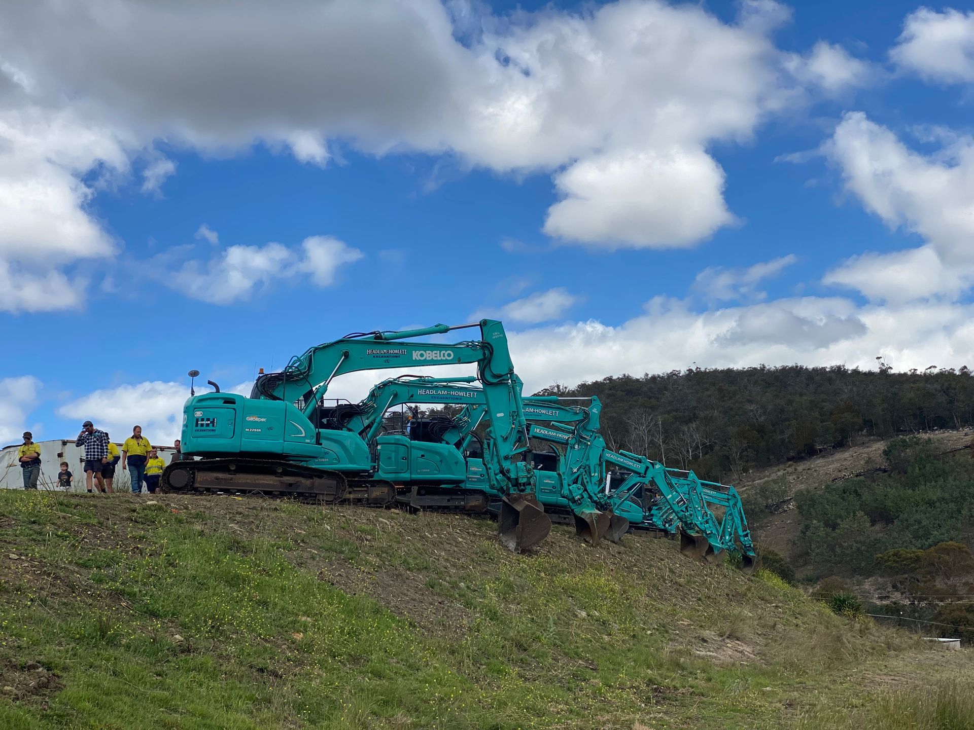 Several green excavators stacked on a grassy hill, with people standing nearby under a cloudy sky.