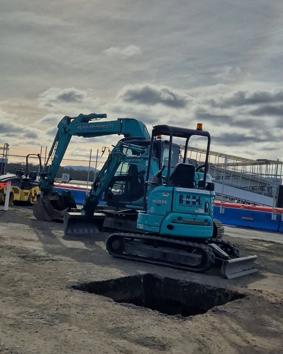 A small teal excavator next to a larger one, in front of a construction site.