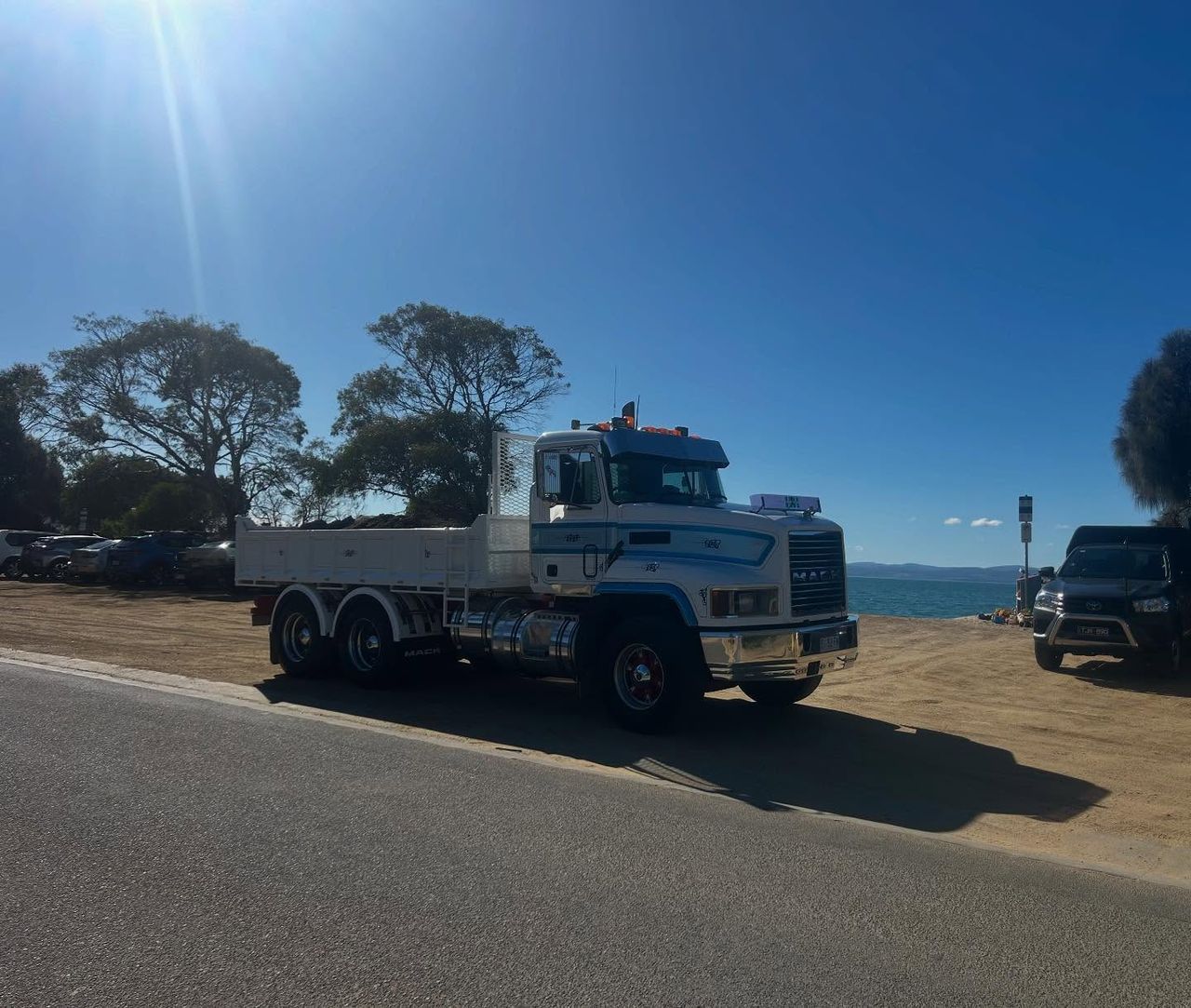 Truck parked near a beach with clear blue sky and trees in the background.