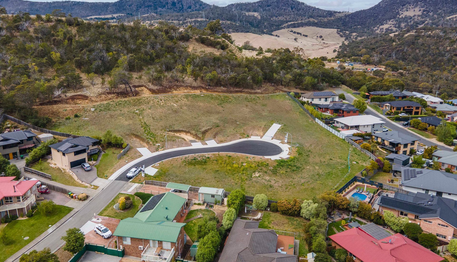 Aerial view of a cleared hilltop with a curved road, surrounded by houses and a backdrop of hills and trees.