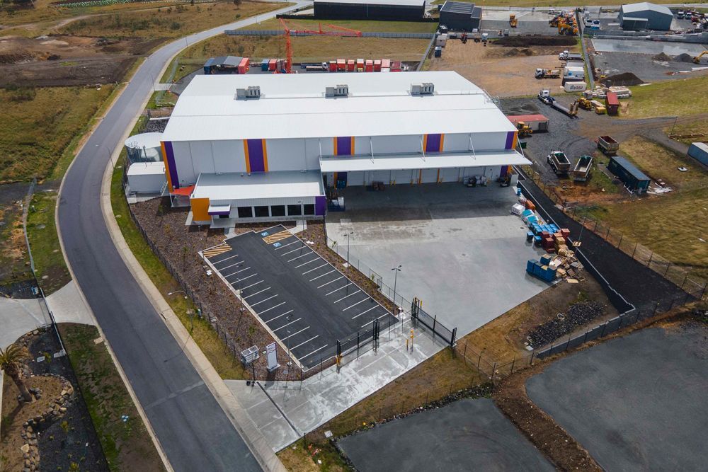 Aerial view of a large warehouse with solar panels in front. The building has a white roof and orange and purple accents.