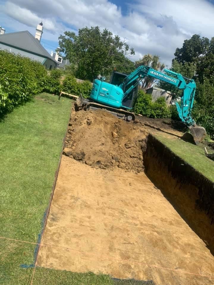 A turquoise excavator digs a rectangular trench in a grassy, sloped yard.