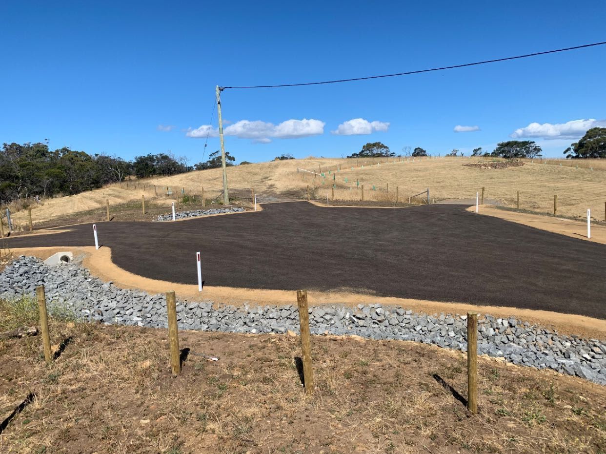 A gravel driveway bordered by rocks and a fence, leading to a grassy hill under a blue sky.
