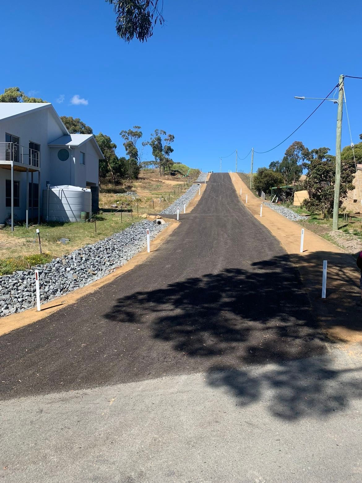 Newly paved road uphill with gravel shoulders, blue sky, and two-story house on the left.