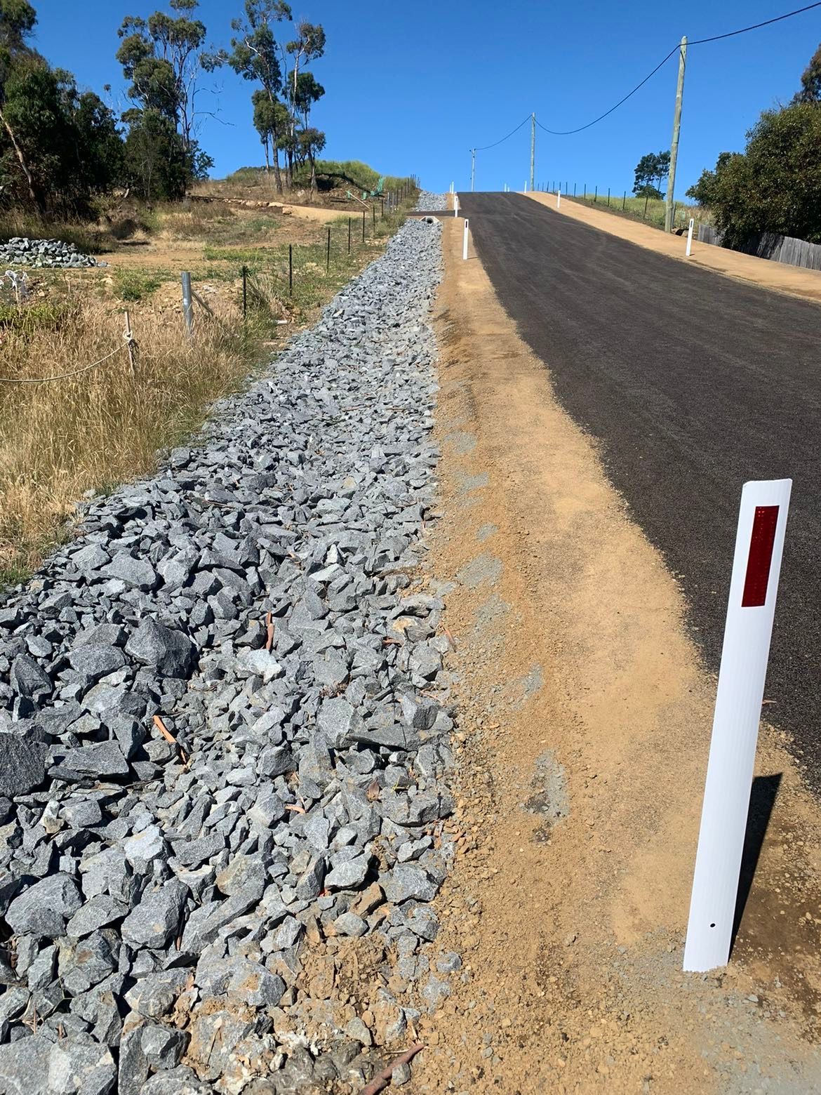Roadside view: New asphalt road bordered by gravel and dirt, ascending a hill.