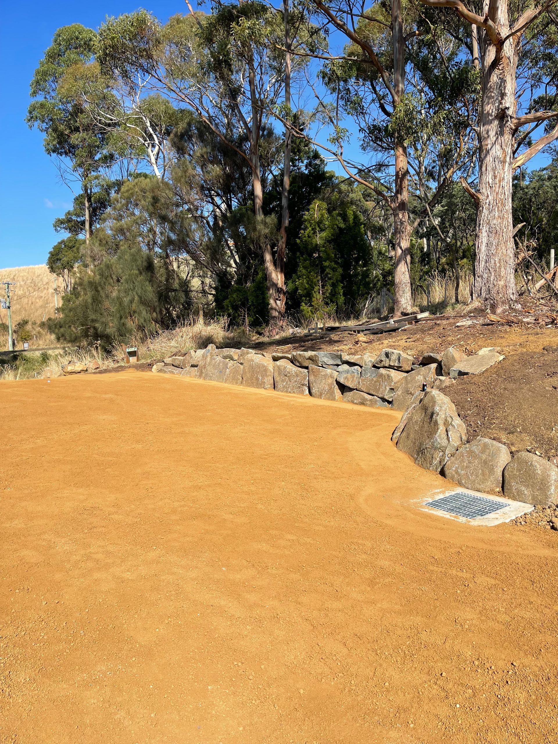 Open area with reddish-brown gravel, bordered by stone wall and trees under a blue sky.