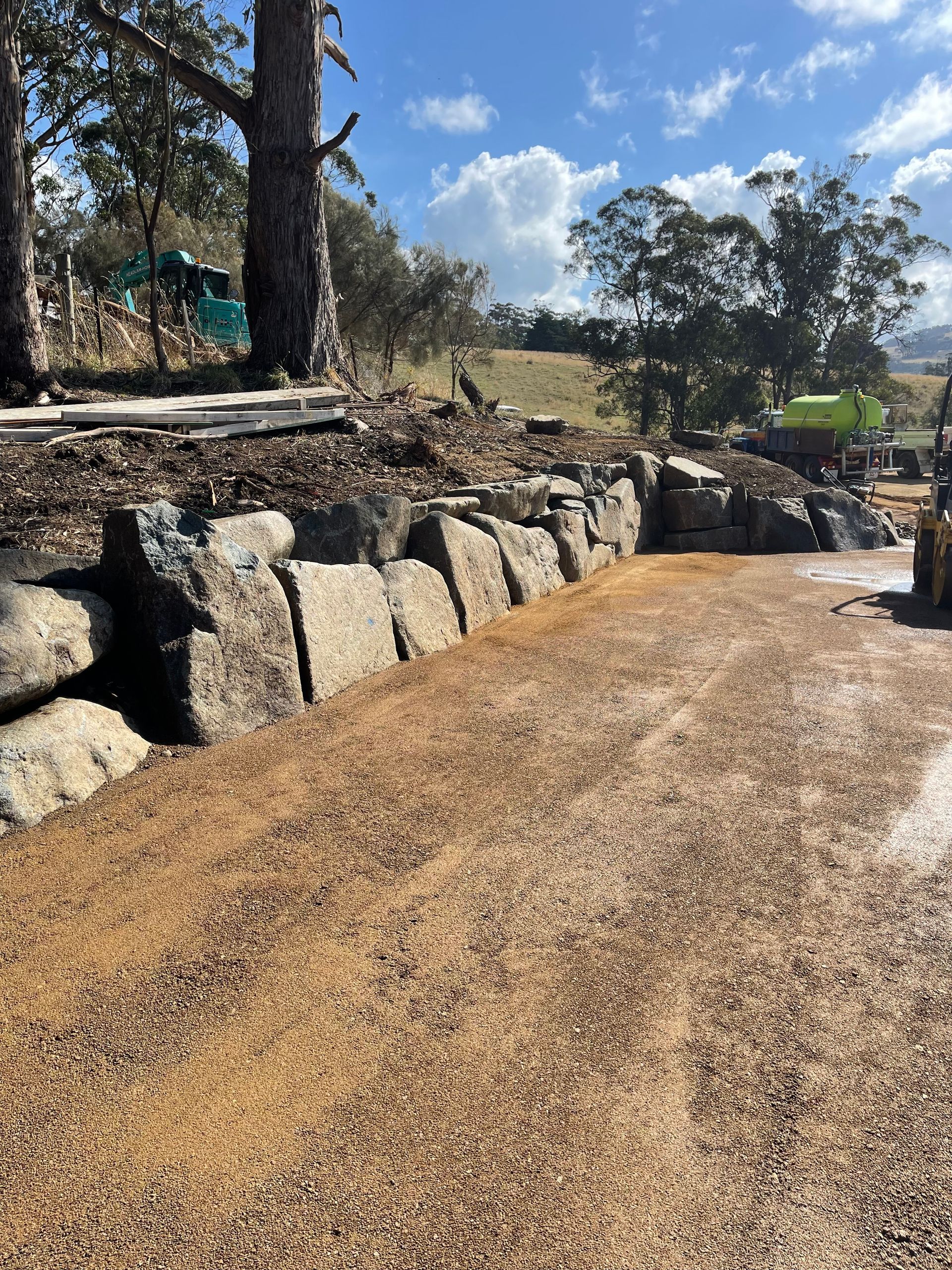 A gravel driveway with a large rock retaining wall under a blue sky.