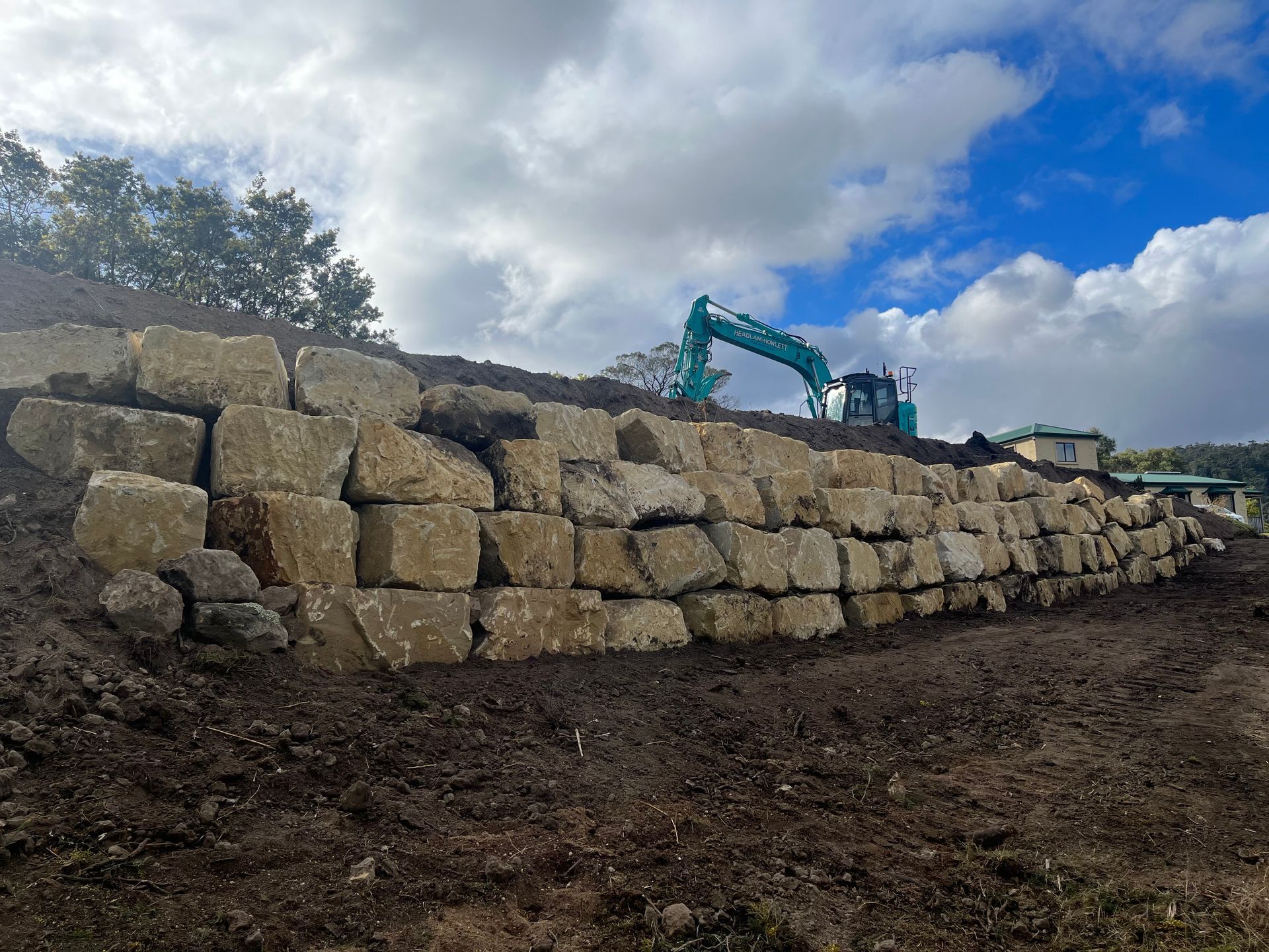 Stone retaining wall on a hillside with an excavator in the background under a cloudy sky.