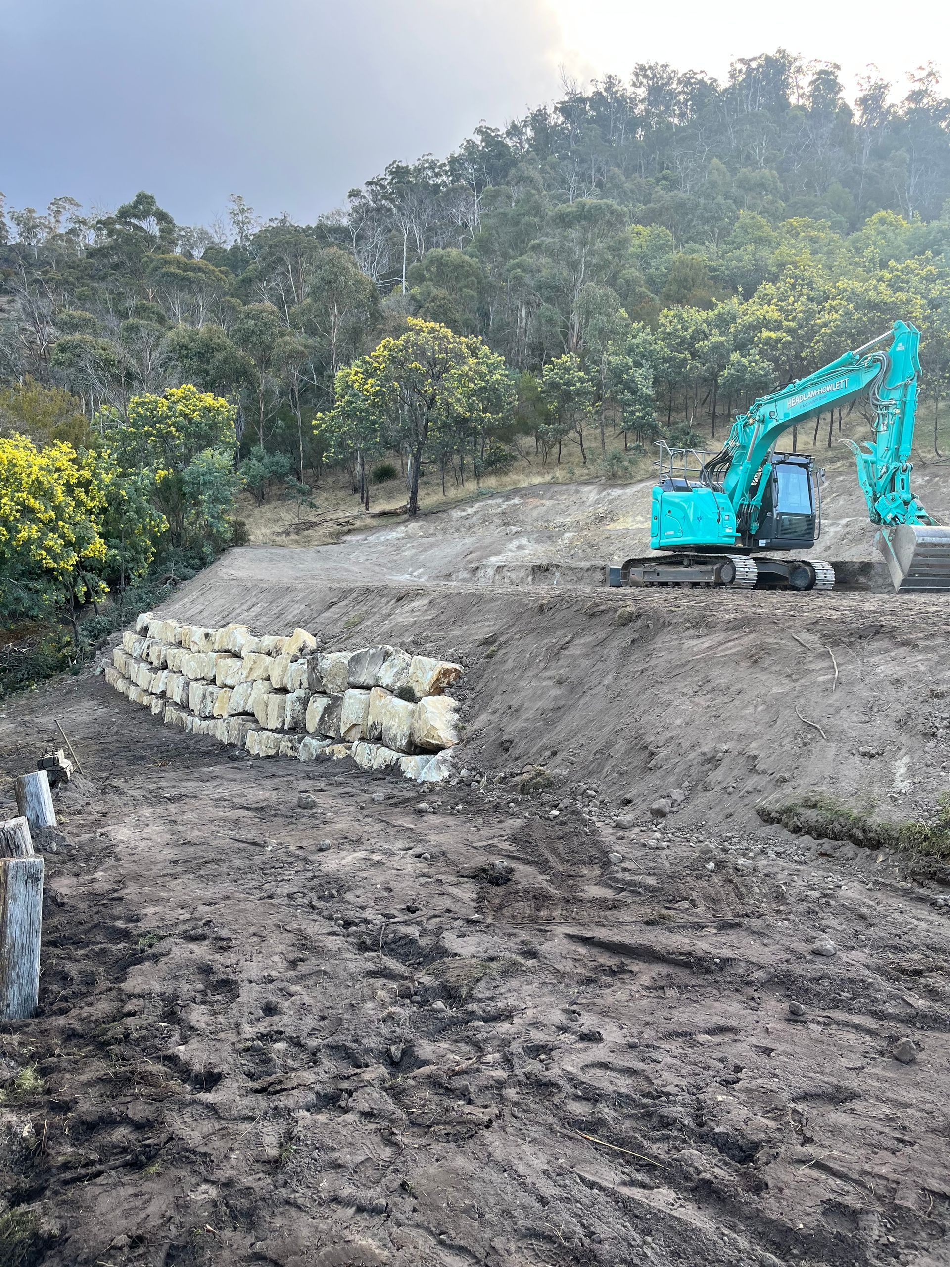 An excavator works on a dirt hillside next to a retaining wall made of large stone blocks.