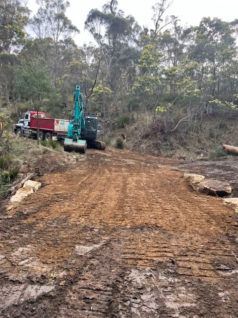 An excavator working on a muddy dirt road next to a red dump truck, surrounded by trees.
