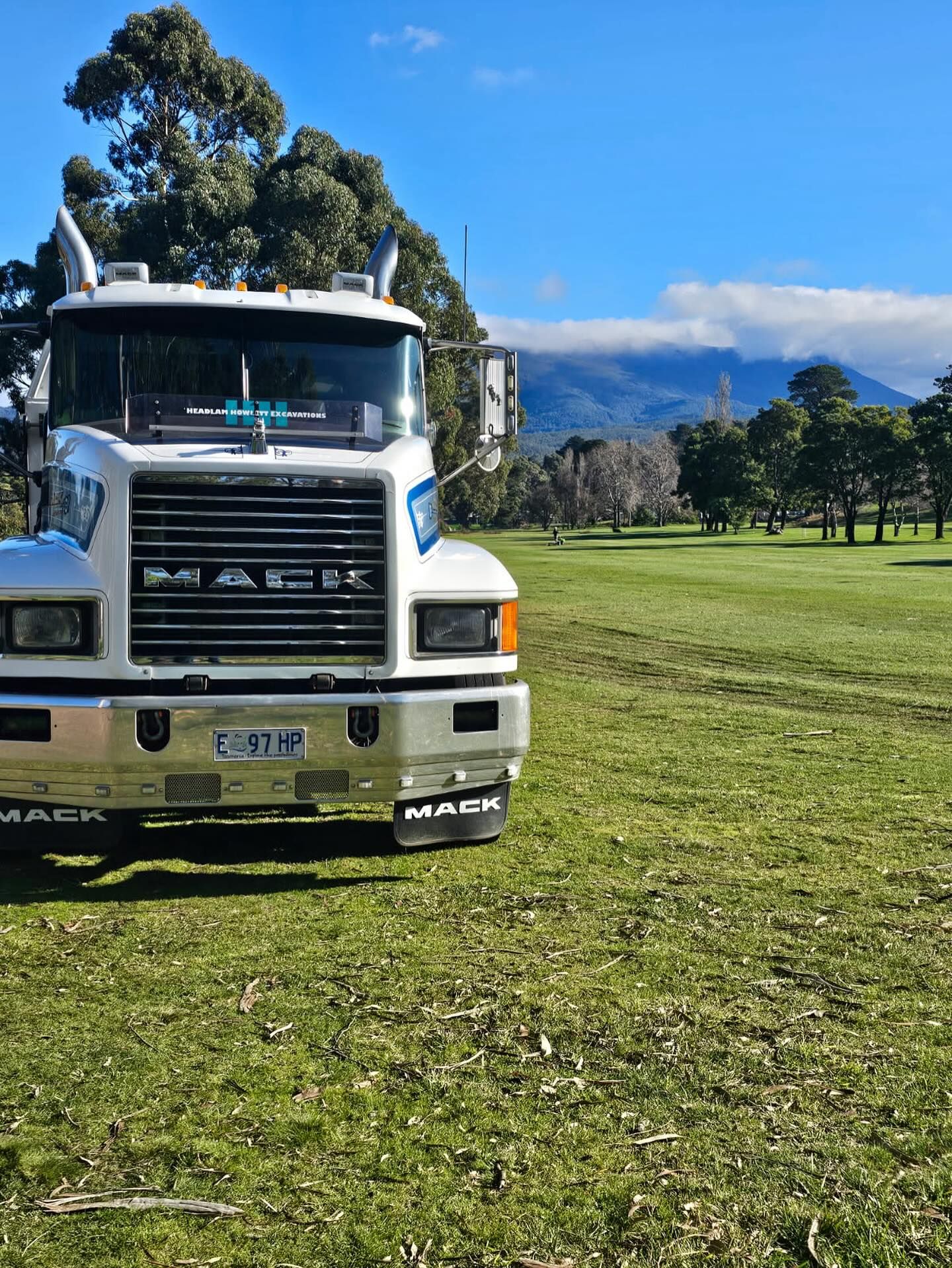 White Mack truck on a grassy field with trees, and a mountain in the background.