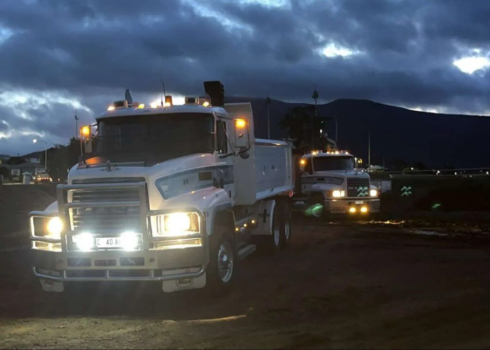 Two white semi-trucks with headlights on at dusk, in front of a dark mountain silhouette.