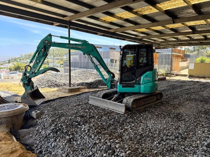 A compact green mini excavator parked under a partially covered structure, positioned on a bed of crushed gravel with construction materials.