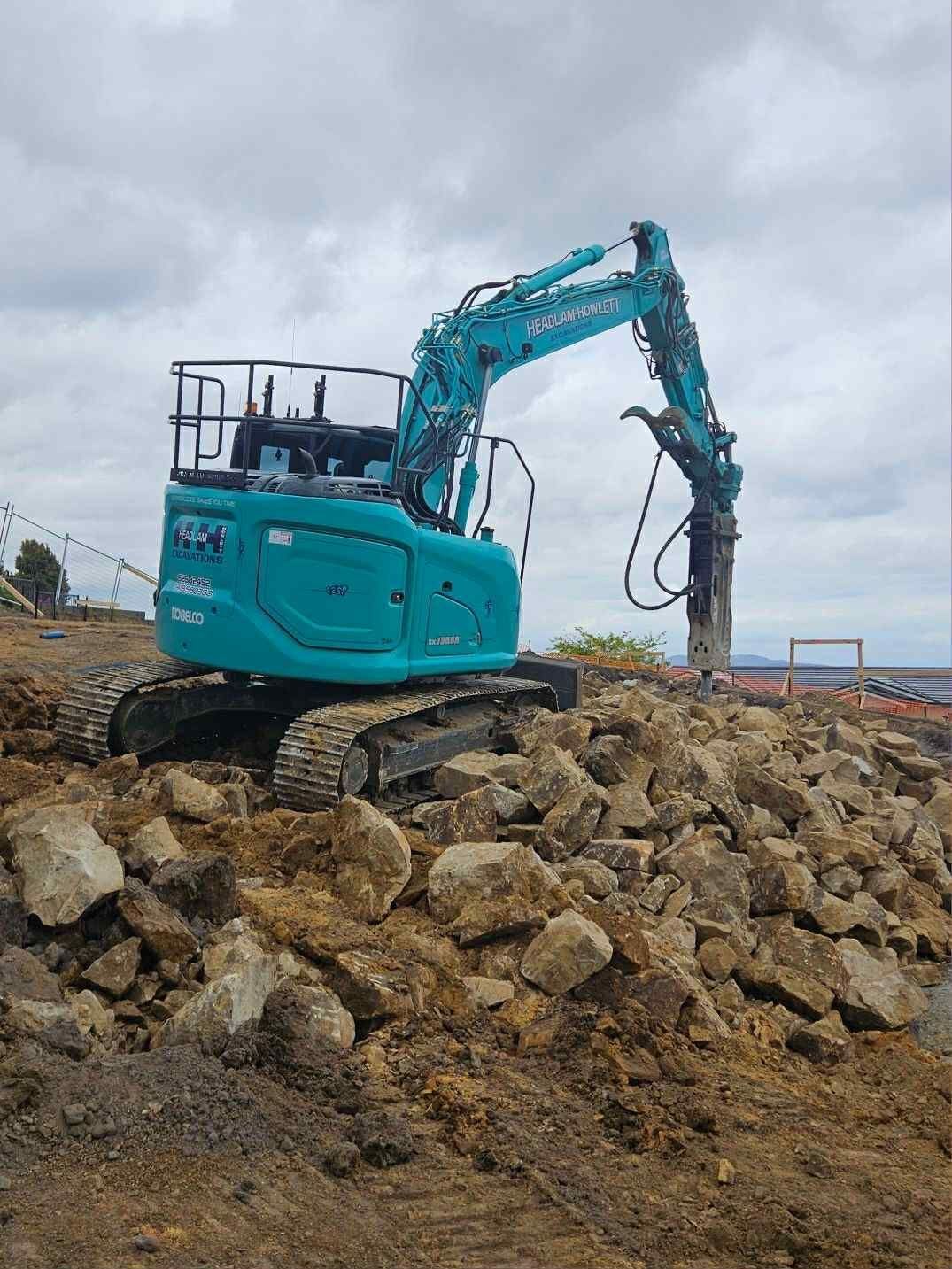 Blue excavator with a jackhammer attachment on a pile of rocks at a construction site.