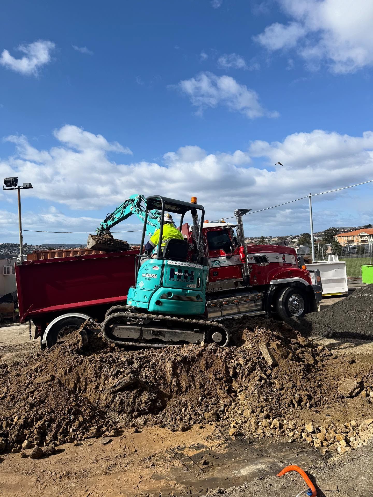 A small excavator loading a red dump truck with dirt on a construction site under a blue sky.