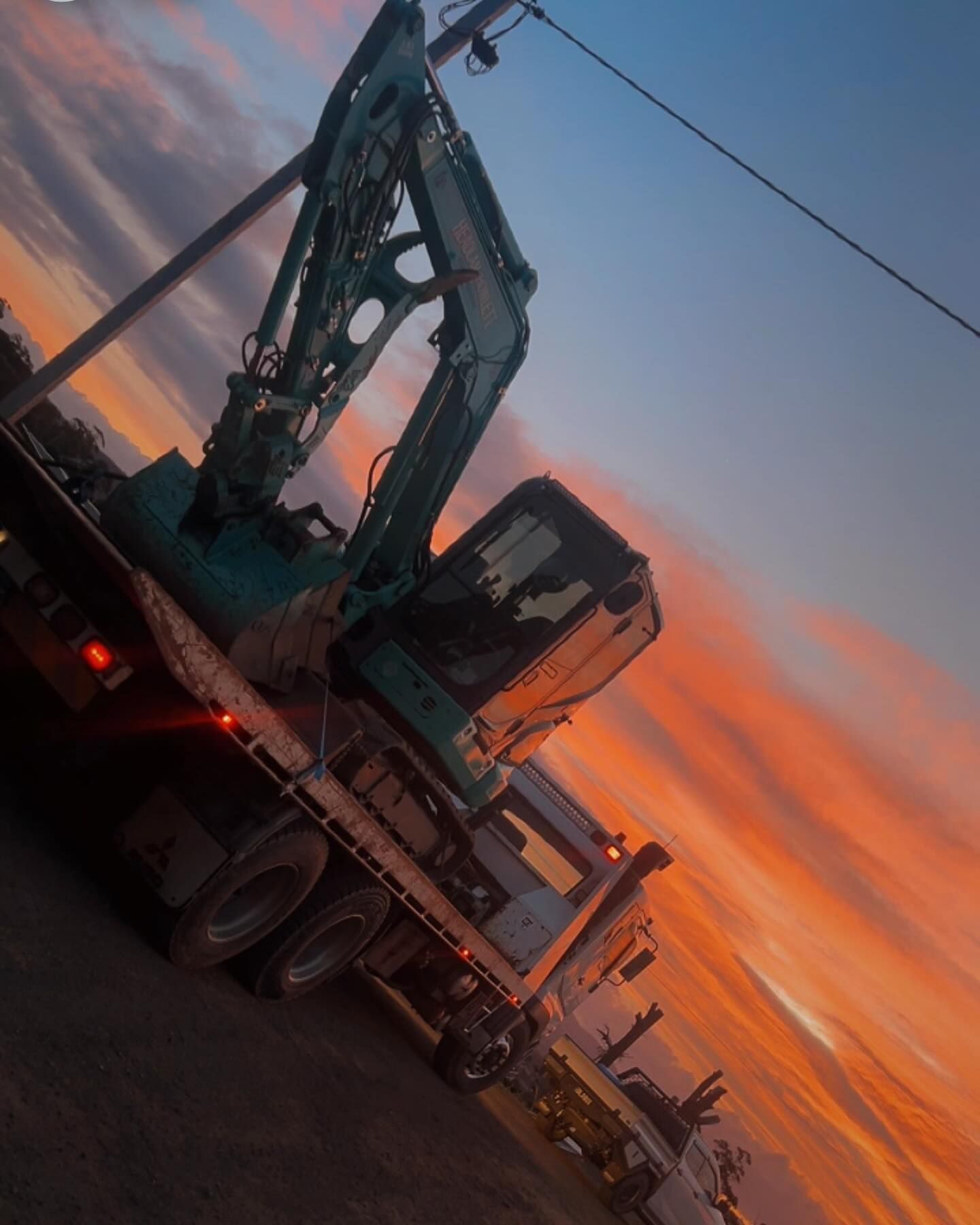 An excavator on a truck bed against a colorful sunset sky.