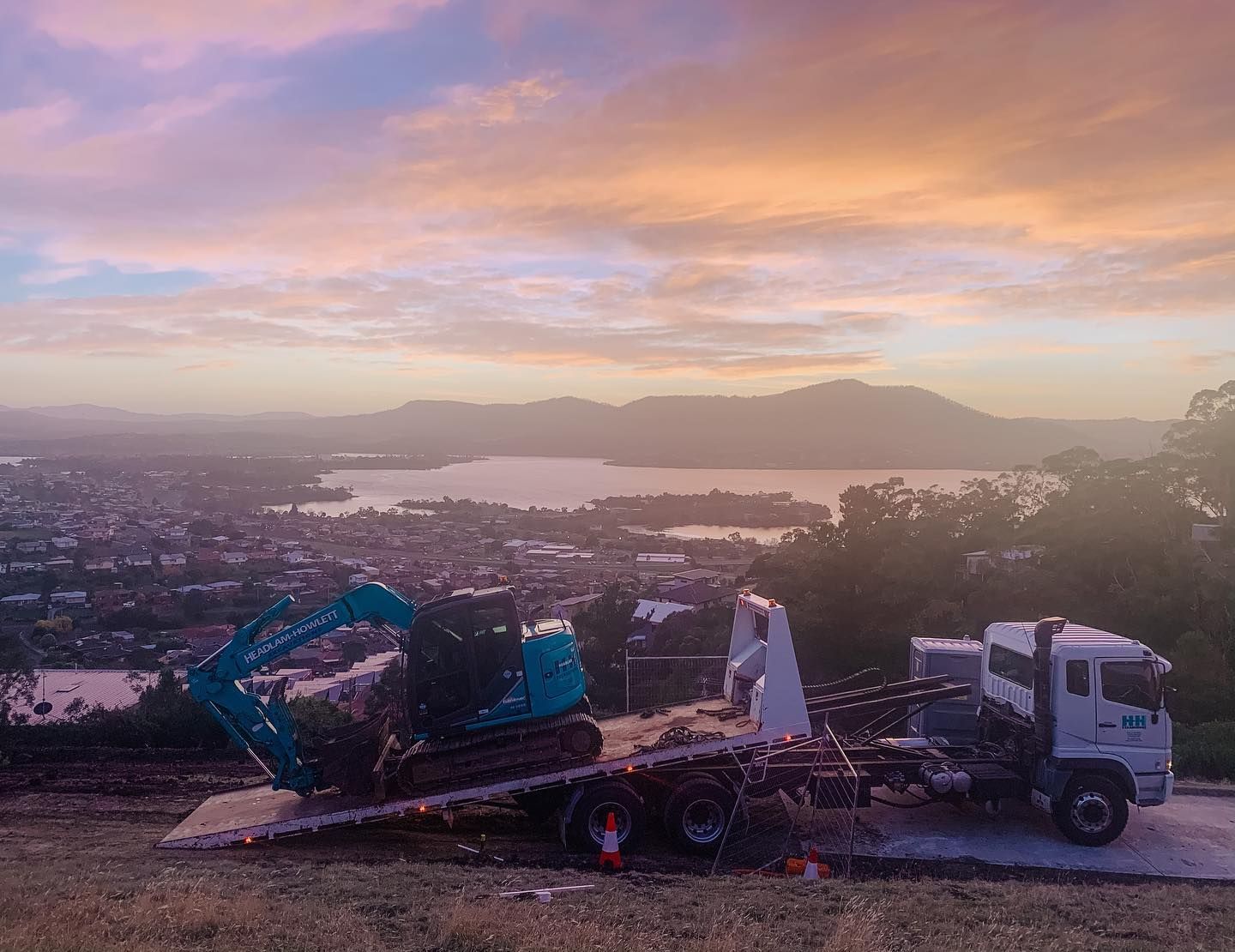 Truck with excavator on flatbed, overlooking a city, lake, and colorful sunset.