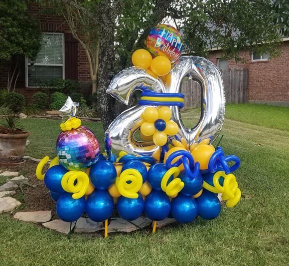 A bunch of blue and yellow balloons are sitting in the grass in front of a house.