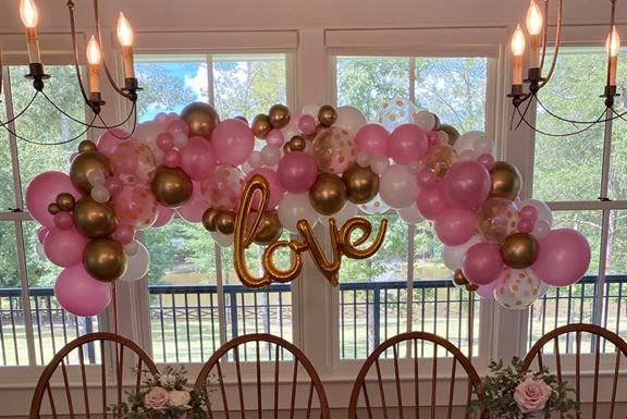 A table and chairs are decorated with pink and gold balloons and a love sign.