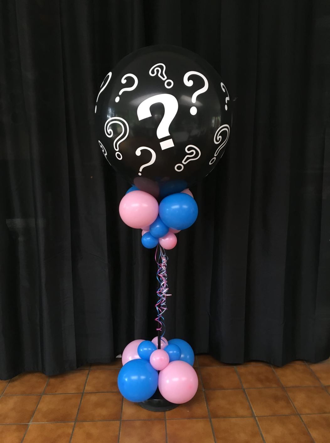 A black balloon with a question mark on it is surrounded by pink and blue balloons.