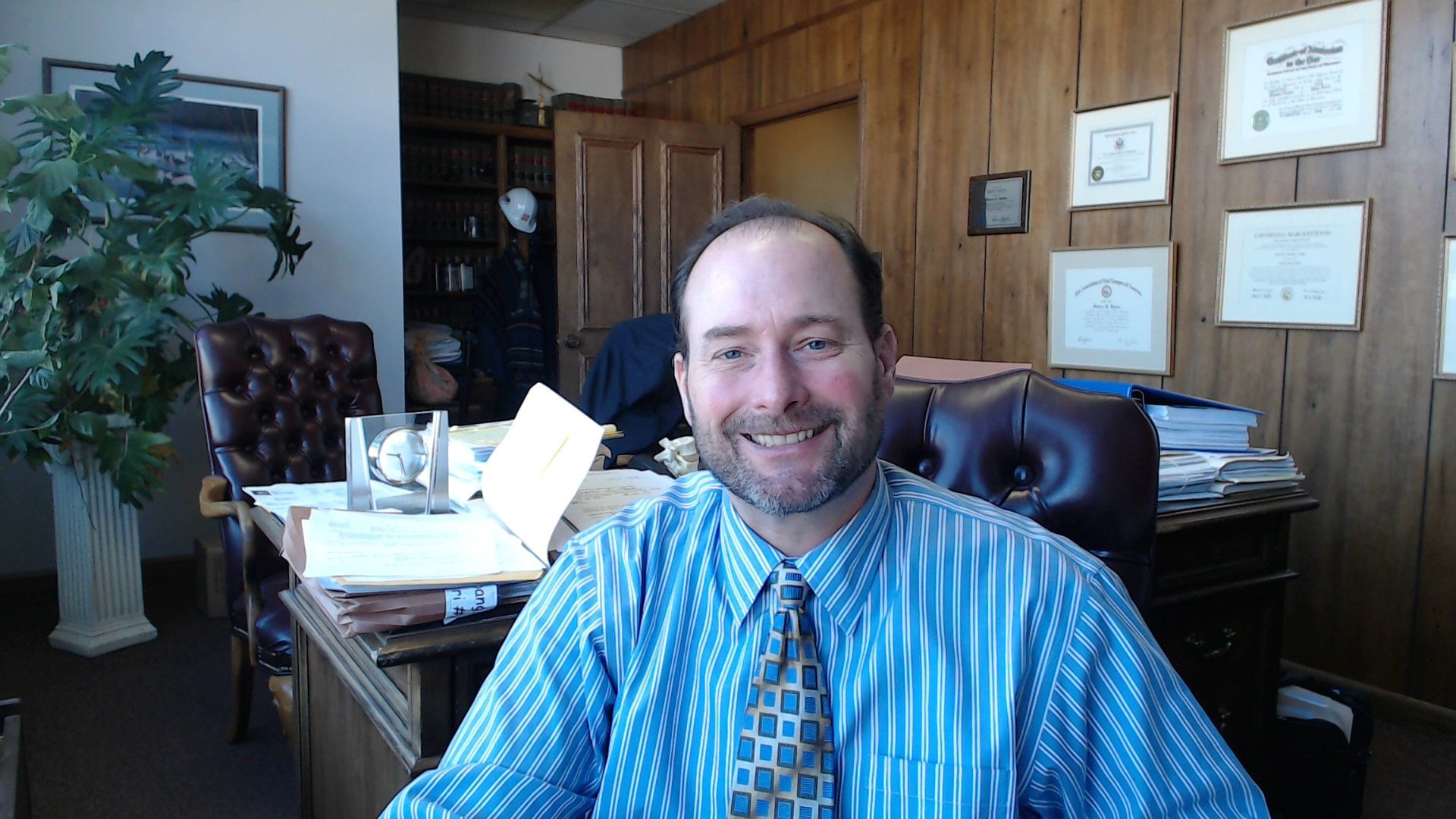 Man in blue striped shirt smiles in a wood-paneled office, papers on desk, certificates on wall.