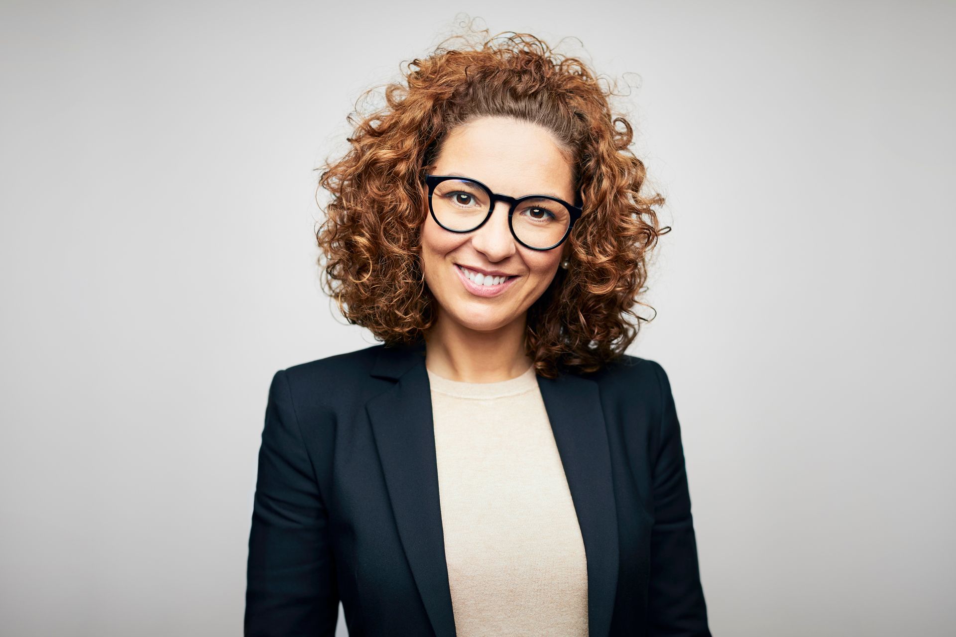 A woman with curly hair and glasses is wearing a suit and smiling.