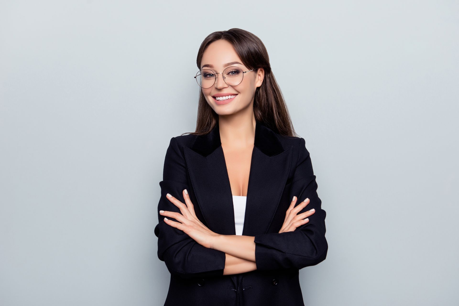 A woman in a suit and glasses is standing with her arms crossed and smiling.