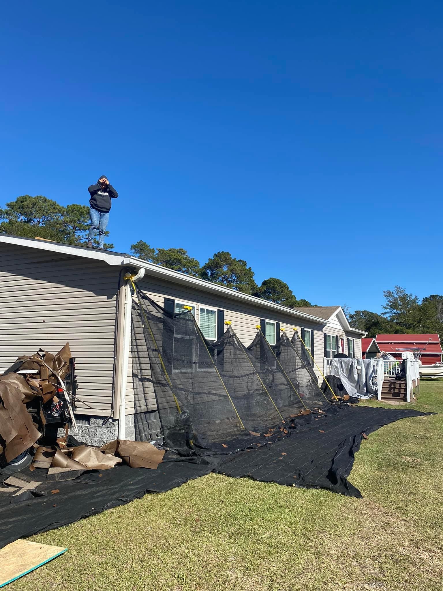Damaged wall beside a mobile home with debris on the ground and a worker on the roof under a blue sky