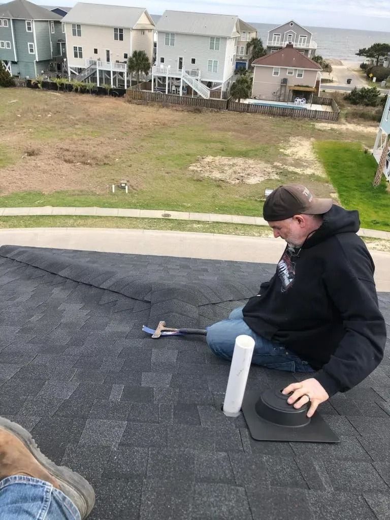 Person sitting on a shingled roof near a white pipe, looking toward a grassy neighborhood below.