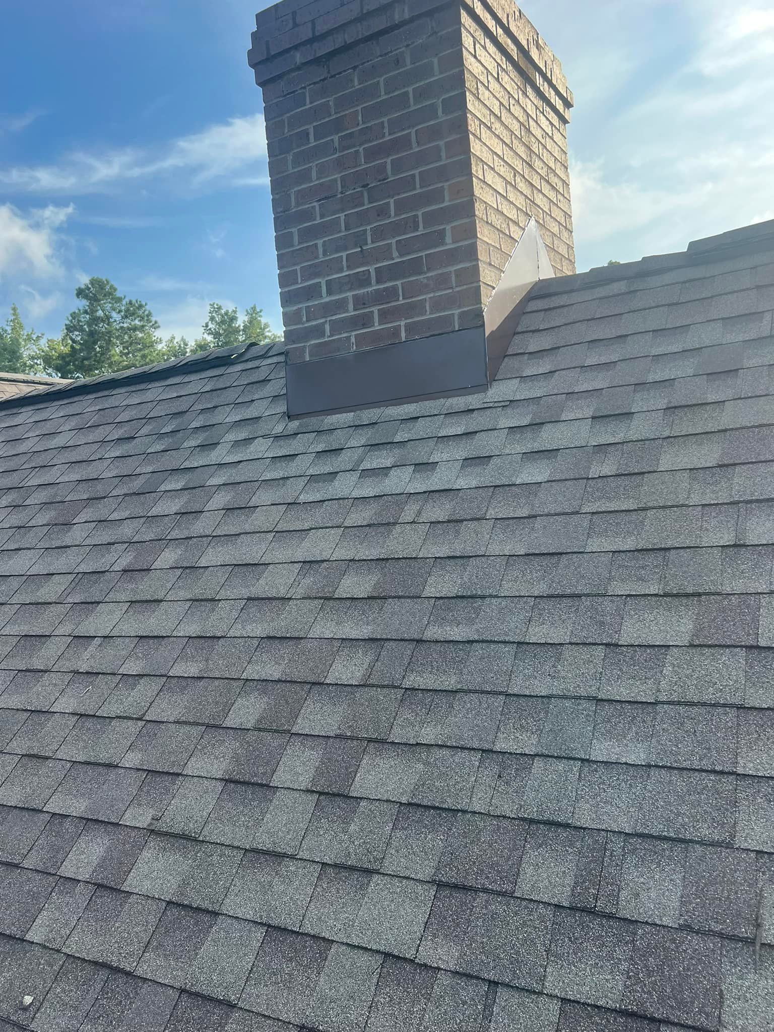 Gray shingled roof with a brick chimney against a blue sky and trees
