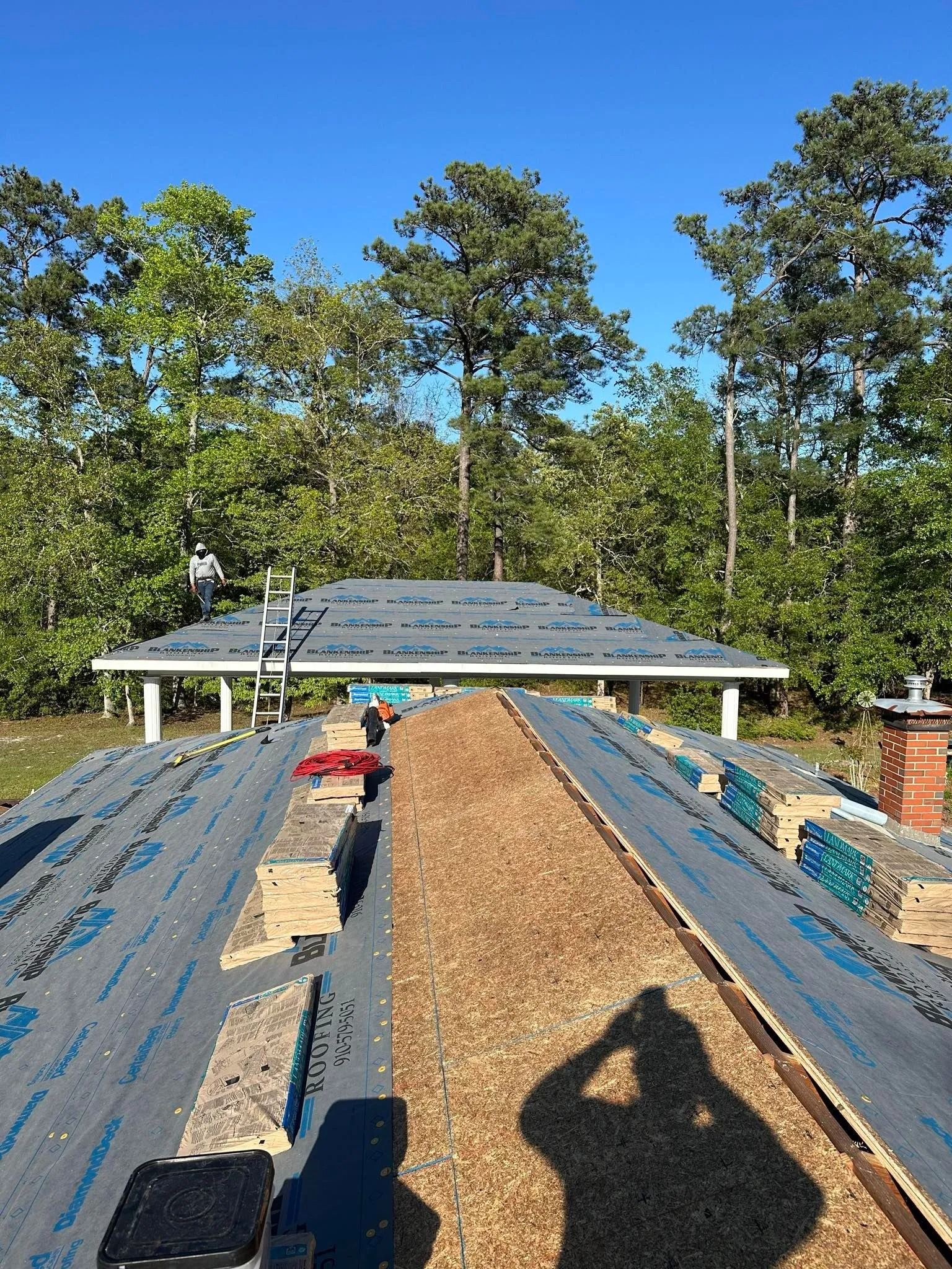 Wooden roof under construction with blue tar paper and wood chips, surrounded by trees on a sunny day