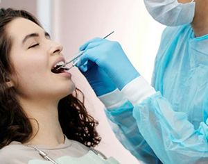 A dentist examining a patient's teeth with tools in a dental office. The patient has their mouth open, eyes closed.