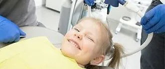 A young girl smiles during a dental visit. She lies in a chair, with gloved hands and equipment present.
