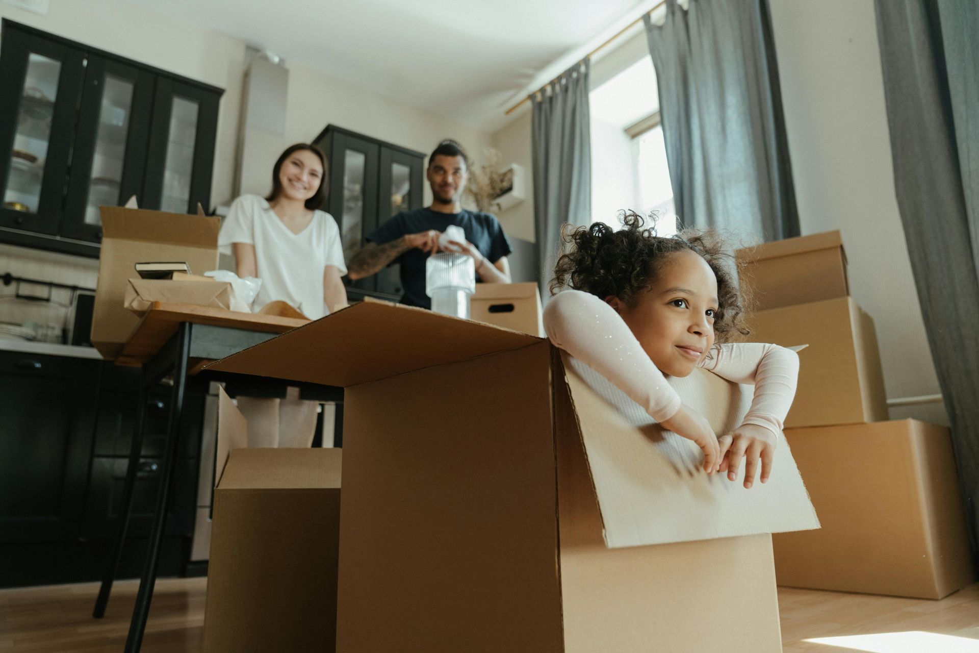 A little girl is sitting in a cardboard box with family in the background.