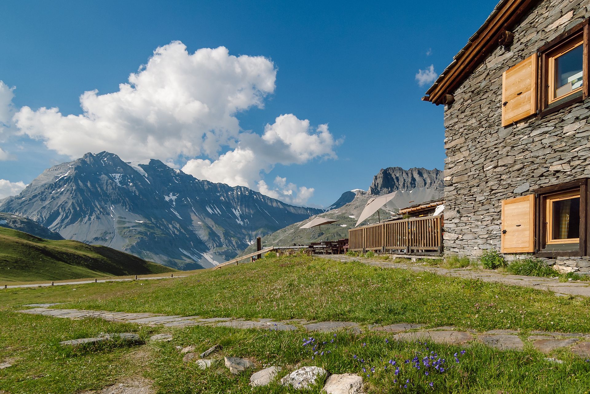 Stone houses in a mountain village beside a grassy meadow and snow-capped peak under a blue sky